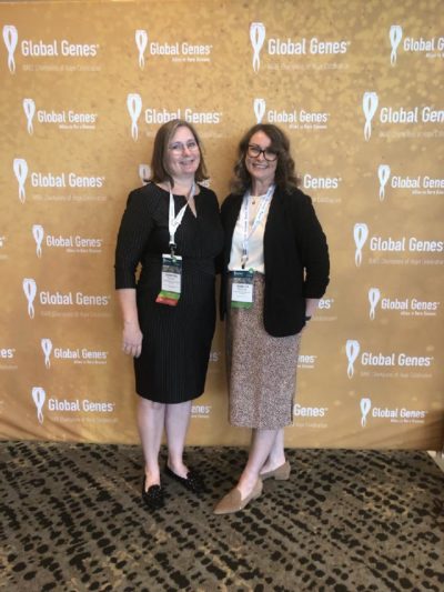 Photo depicts founding members, Christina and Rebecca, standing in front of a Global Genes gold colored back drop.  Both women are dressed in business casual clothing and smiling.
